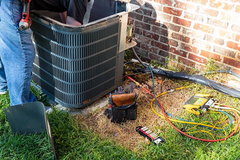 Photo of an air conditioning unit installed outside of a brick home being worked on by a technician.