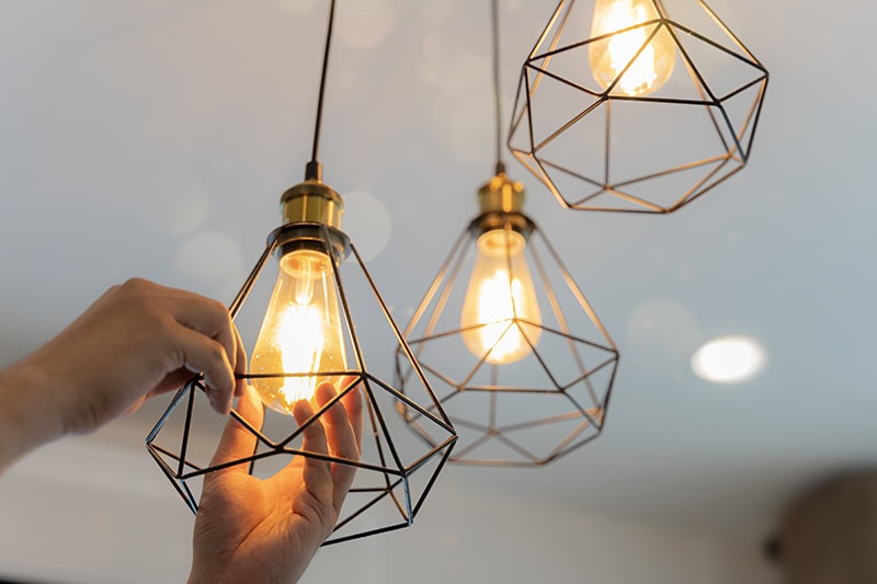 A close-up shows a person's hands carefully installing or adjusting a glowing Edison-style LED light bulb inside a modern, geometric black wireframe pendant cage.